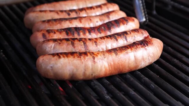 A close-up shot of delicious, juicy bratwurst sausages cooking on a hot charcoal grill. The sausages feature perfect char marks and a glistening texture, representing a classic summer outdoor barbecue
