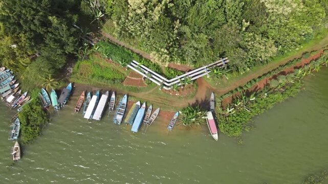 A high-angle drone shot moves steadily to the left, revealing a row of colorful traditional passenger boats moored along the grassy banks of Gajah Mungkur Reservoir
