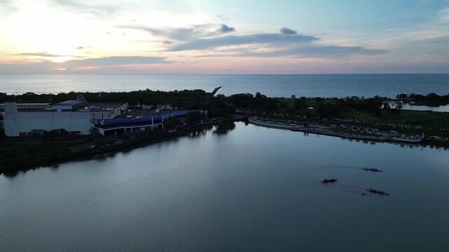 A smooth pedestal drone shot rises vertically to reveal traditional rowing boats gliding across the calm waters of Tanjung Bunga during a serene sunset