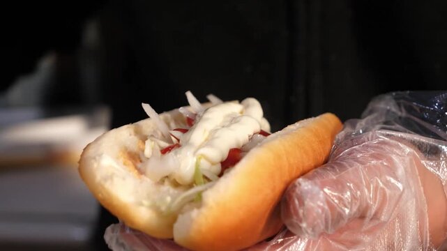 Close-up of a food vendor wearing plastic gloves preparing a delicious hot dog. The footage shows a white condiment, likely mayonnaise or a creamy sauce, being squeezed from a plastic bottle onto a ho