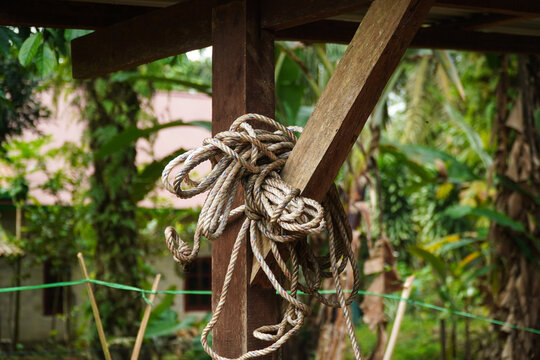 Coiled thick rope hangs loosely from a weathered wooden post in a lush green outdoor setting with dense foliage and natural light filtering through leaves creating dappled shadows on the ground.