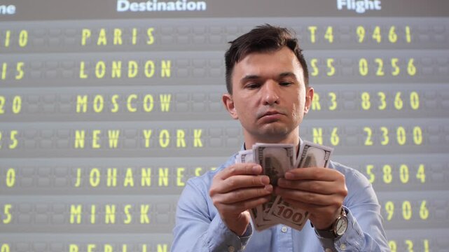 Man holding american dollar banknotes, calculating travel expenses and budgeting for a trip with international destinations displayed on the airport departure information screen