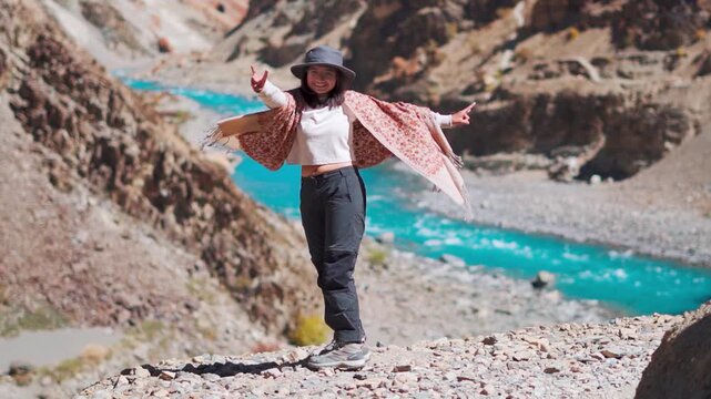 Happy Indian teenager girl wearing cap and enjoying trip in mountains, tourist admiring beauty of nature. Girl standing near blue Tsarap river in Zanskar, Ladakh, India. Travel concept.