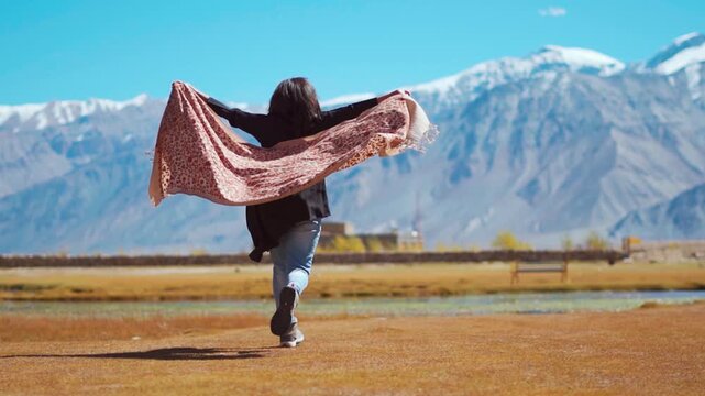 Happy Indian woman running in meadow in front of Himalaya mountains in Sani village, Zanskar, India. Woman holding shawl in hand. The concept of achieving success. Happy and drunk on life.
