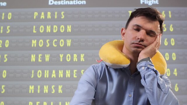 Man with neck pillow asleep in busy airport terminal, resting against chair while departure board shows delays and cancellations during a long layover, travel fatigue visible