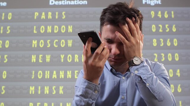 Angry passenger expressing his frustration with travel disruption and flight delays, talking loudly on his mobile phone near an airport information display