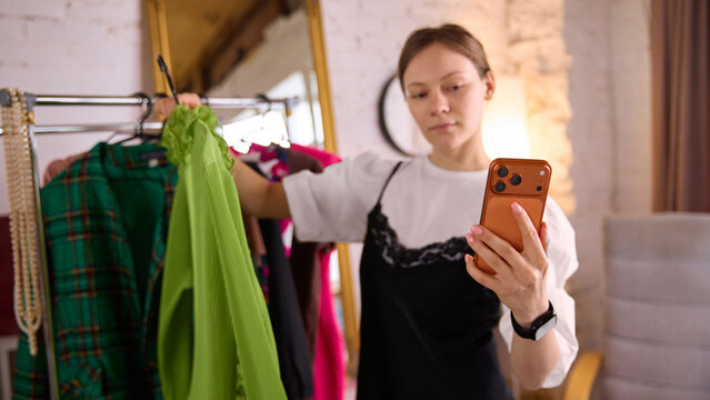 Woman choosing green dress while browsing smartphone at home. Concept of fashion decision process, online shopping integration, product evaluation, consumer choice behavior