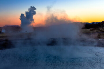 Fototapeta premium Sunset image of a geyser eruption, silhouetting a distant, indistinct figure showing scale amid a steamy landscape, Strokkur Geyser, Iceland 