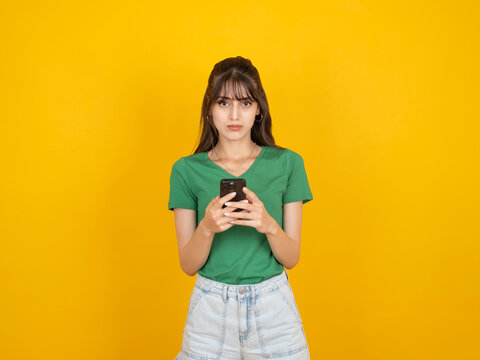 Serious caucasian woman holding smartphone and looking at screen with focused expression, reading message or content, wearing green t-shirt and denim, isolated on yellow background with copy space.