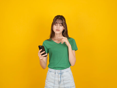 Thoughtful caucasian woman holding smartphone and touching chin with pensive expression, considering message or decision, wearing green t-shirt and denim, isolated yellow background with copy space.
