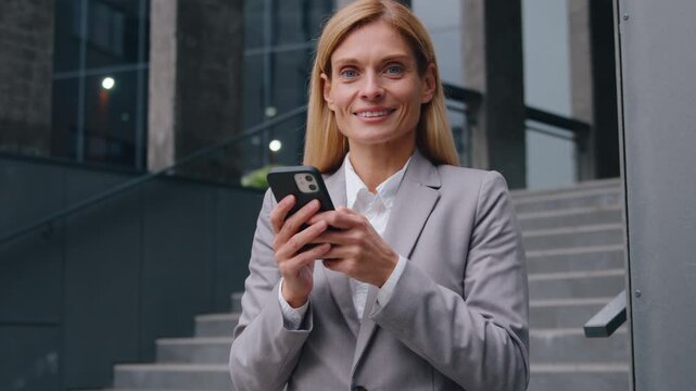Satisfied Caucasian middle-aged business woman holding mobile phone smiling looking at camera happy adult businesswoman portrait with smartphone device in downtown female using internet app technology