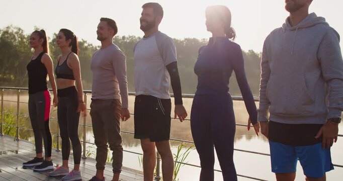 Group of young adults active women and men dressed in sportive wear performing squats together standing in row on boardwalk near lake during sunrise. Fitness, and healthy lifestyle on nature