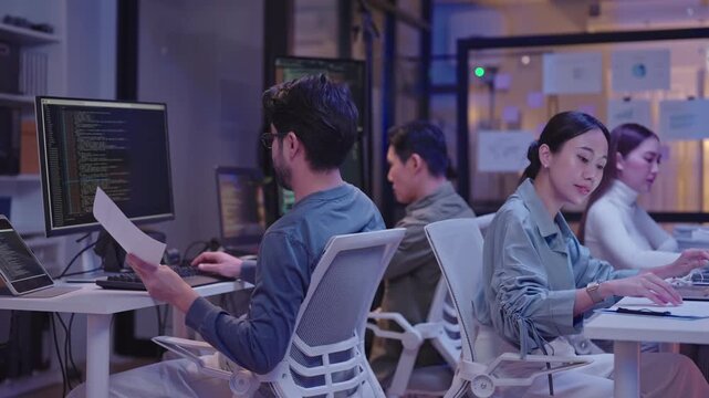 Side view of focused Asian male professional at desk receiving a paper document from an unseen colleague, then quickly turning back to continue working on his laptop during a late-night office shift.