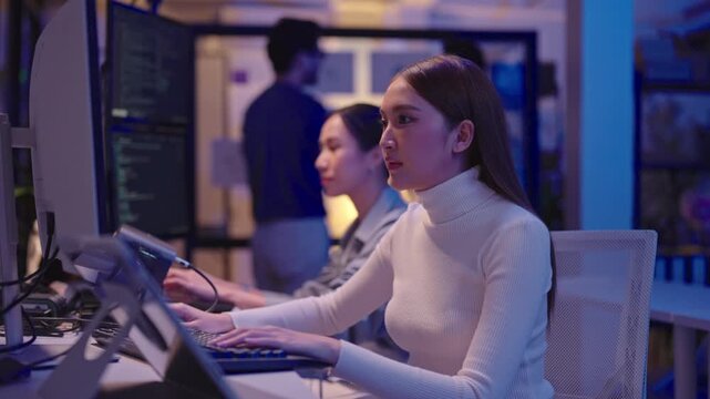 Focused Asian programmers working on multiple screens, writing code and developing software during late-night shift in a dark modern office.