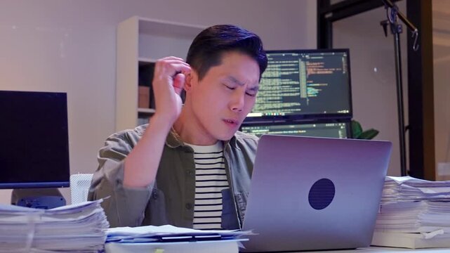 Mature Asian professional man looking serious while working on a laptop surrounded by large stacks of documents in a late-night office setting.