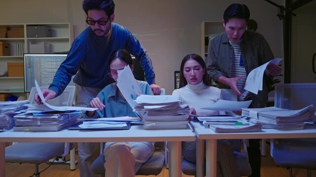 Group of Asian colleagues pan shot searching through disorganized piles of documents, feeling rushed and stressed to meet a night deadline.