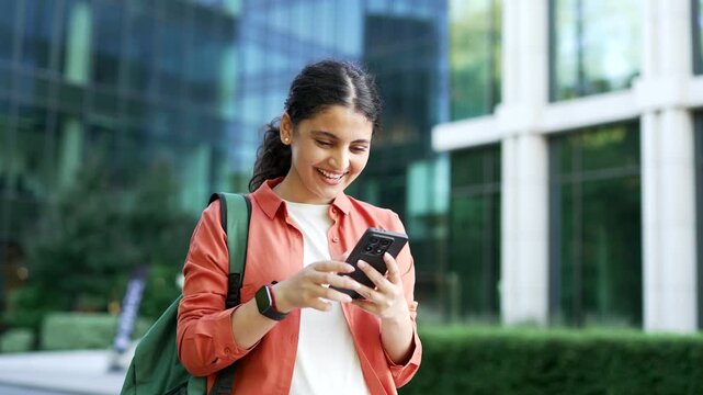 Smiling female student reads a message on her smartphone while standing near university building. Young woman shows pleasant surprise and joy after receiving good news in outdoor academic environment.