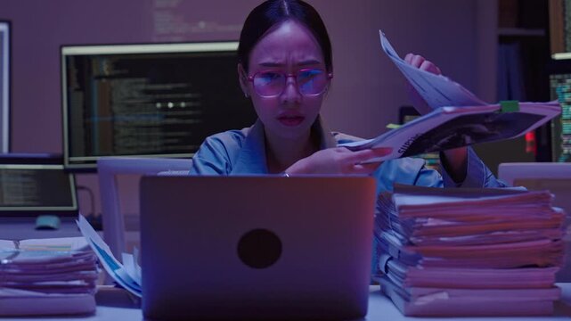 Close up of a frustrated Asian female employee rubbing her eyes and forehead, feeling burned out while searching through documents late at night.