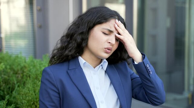Close up of a businesswoman massaging her temples and forehead with hand due to headache near an office building. Female employee shows tension and fatigue, feeling unwell outdoors in an urban setting