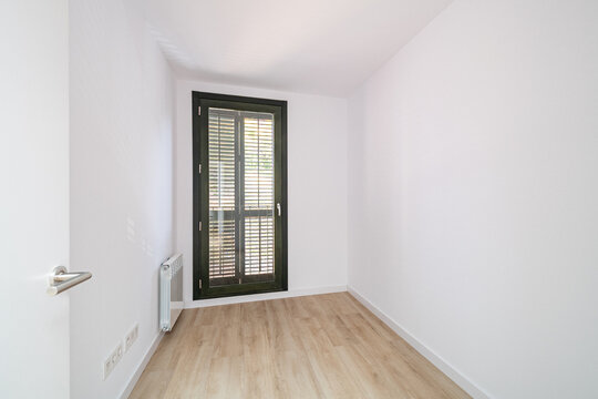 Empty room with white walls and wooden floor. A large window with black frames allows natural light to enter. Minimalist design with no furniture.