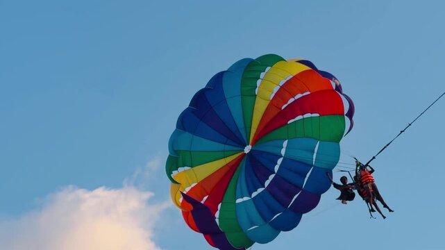 Parasailing activity over the sea at sunset on Phuket island, Thailand. Tourist flying with colorful parachute above the ocean with boats and jet skis on the shore during golden hour.