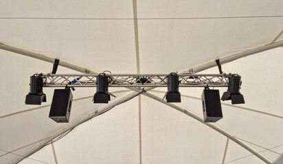 A row of black professional spotlights and audio speakers hanging from a metal truss system against the white canvas background of a large event tent © nahhan