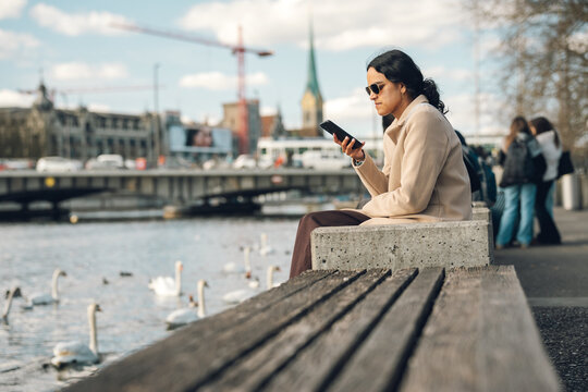 Woman sitting by the limmat river, checking her mobile phone in zurich