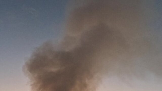 A dense dark smoke plume rises into the open sky, then curls, spreads and reshapes with the wind, creating a dramatic atmospheric background with drifting haze and changing density at dusk.