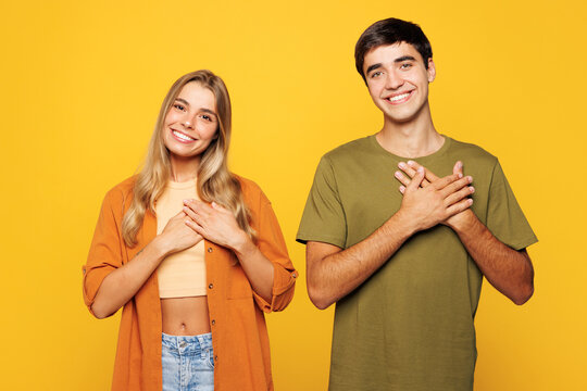 Young calm smiling thankful grateful couple two friends family man woman they wear casual clothes together put folded hands on hearts look camera isolated on plain yellow background studio portrait