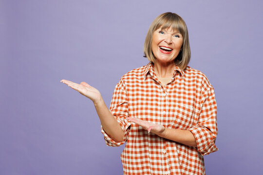 Elderly blonde happy woman 50s years old wear beige t-shirt shirt casual clothes point index finger aside indicate on area mock up isolated on plain pastel light purple background. Lifestyle concept.