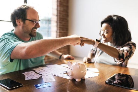 Two people fist bump over table with a piggy bank, teamwork and savings. Diverse man and woman fist bump on financial planning. Couple fist bump teamwork on financial, accounting and investment topic