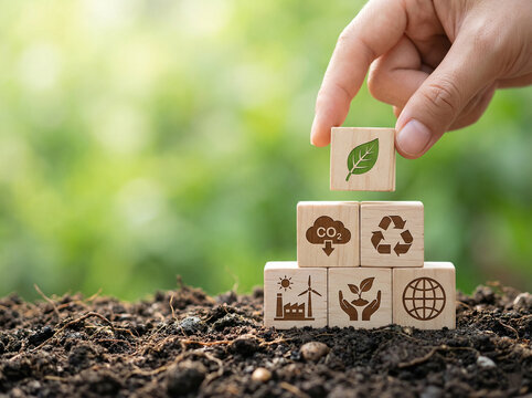 wooden blocks with environmental icons stacked on soil with hand placing top block symbolizing sustainable growth and corporate esg strategy