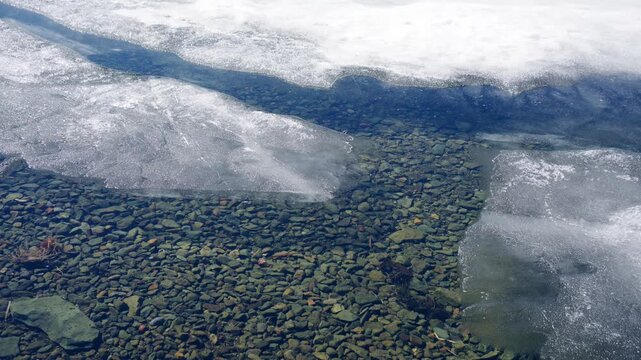 Melting ice on the shore of lake in early spring. Ice floes floating on the water. Colorful stones on the bottom of lake. Beautiful spring nature
