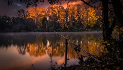 Fototapete Reflection Fishing rod rests along still water's edge, with golden trees reflected beneath a dramatic, cloudy sky  © mbk