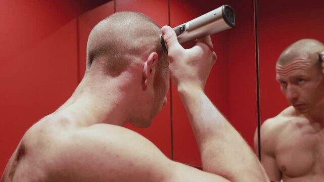 Man shaving his head at home using electric hair clipper in bathroom mirror. Self grooming and personal care routine.