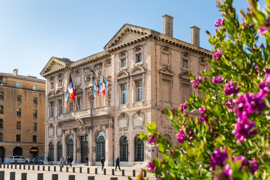 H&ocirc;tel de Ville de Marseille vue de trois quart avant, fa&ccedil;ade principale avec drapeaux, plusieurs personnes qui marchent sur le parvis, arbuste fleuri en premier plan &agrave; droite de l'image