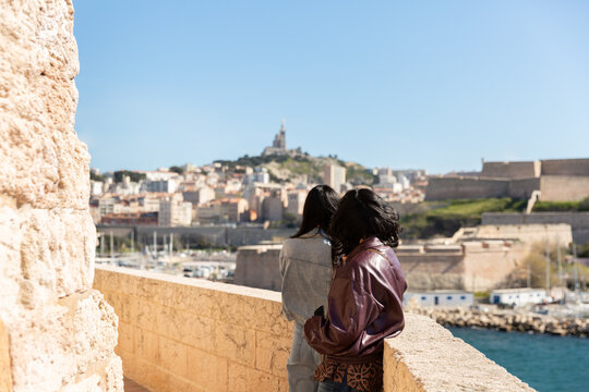 Deux touristes femmes vues de dos debout contre un muret surplombant la sortie du Vieux-Port de Marseille regardant la ville, Notre-Dame de la Garde en arri&egrave;re-plan, soleil et ciel bleu, Sud de France