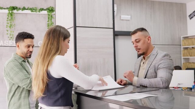 Professional consultant assisting a young couple at a modern office reception. The advisor explains documents and shows a catalog to the clients