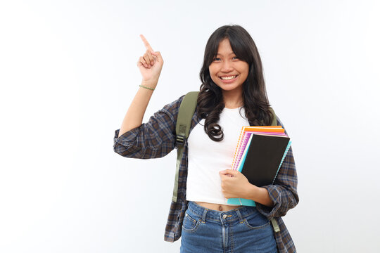 Expressive Asian teenage girl wearing backpack and carrying books, pointing up at empty space, standing isolated on grey background.