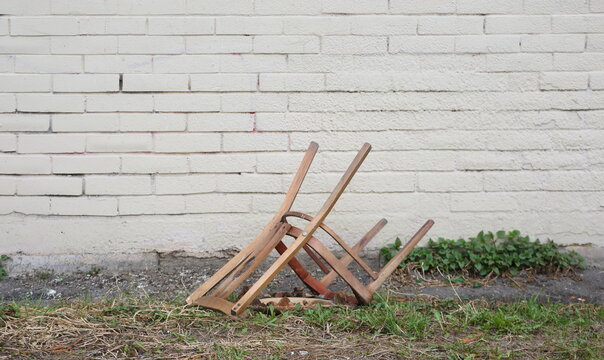 A broken wooden chair lies on the grass near a brick wall
