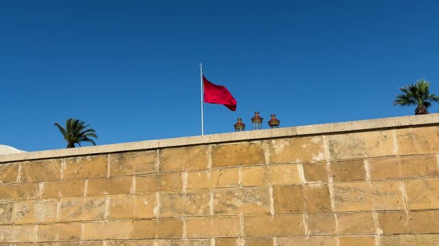Moroccan flag waving on a tall flagpole against a clear blue sky. National symbol of Morocco fluttering in the wind in Rabat city. Tropical landscape featuring palm trees and traditional street lamps.
