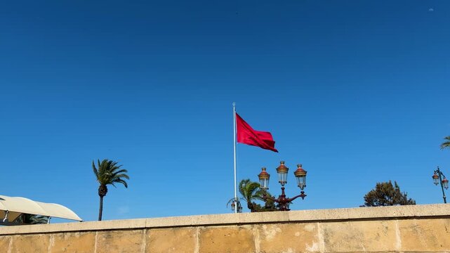 Moroccan flag waving on a tall flagpole against a clear blue sky. National symbol of Morocco fluttering in the wind in Rabat city. Tropical landscape featuring palm trees and traditional street lamps.