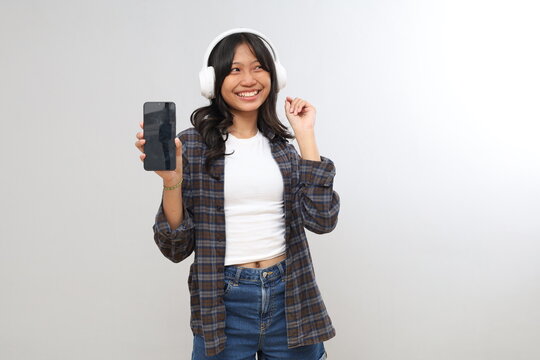 A young Asian teenage girl shows a blank screen of her smartphone while listening to music through headphones, isolated on a gray background.