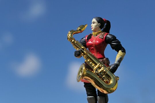 GI JOE action figure of asian female intelligence agent and martial arts specialist Jinx holding tenor saxophone, blue skies with some clouds in background. 