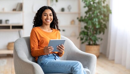 Obraz premium A smiling woman with curly hair sits in a gray armchair, holding a tablet. She wears an orange shirt and jeans in a bright living space