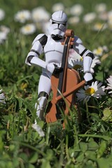 Naklejka premium Action figure of clone trooper from Star Wars movies, in white armor and helmet playing on model of violoncello in garden lawn with blossoming common daisies, sunlit by morning sunshine. 
