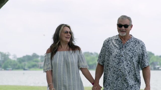 An older man and woman walk along the water's edge at a lake as they hold hands and smile.
