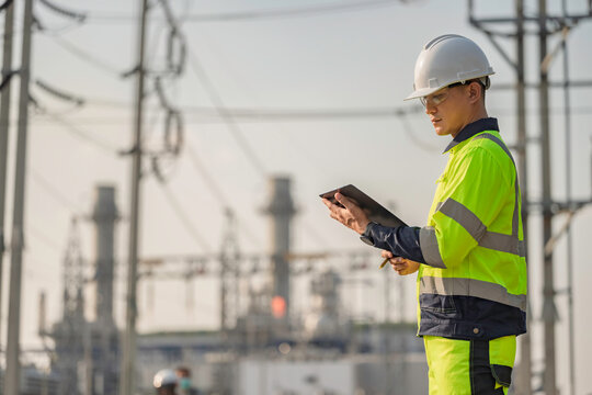 Asian male electrical engineer holding digital tablet standing at power plant station. Professional energy industry technician working on infrastructure maintenance and renewable resource project.