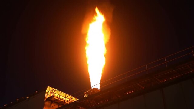 Static shot of industrial flare stack burning and emitting large flame on rooftop refinery platform at night