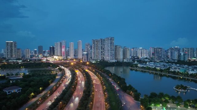 Wide aerial panorama of curving boulevard in Hanoi with continuous traffic streams, modern skyline, lakes, and dense residential districts under blue evening sky